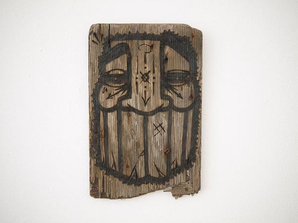 A GATS driftwood sculpture features a carved and painted abstract face with closed eyes and a broad, stylized beard. Its rough texture and faded paint add an aged, rustic charm, set against a plain white background.