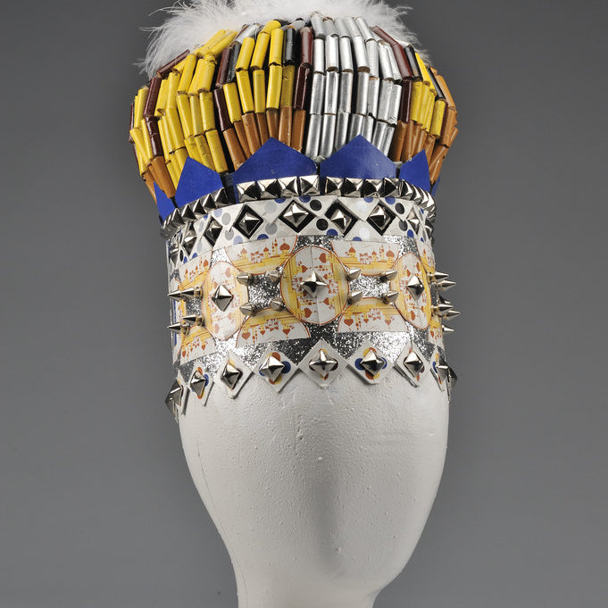 Lucien Shapiros abstract sculpture features a white vase-like base with a mixed-media upper section of colorful, geometric patterns on metallic materials. It is topped with a circular crown-like ornament, sports a feathery texture, and stands against a gray backdrop.