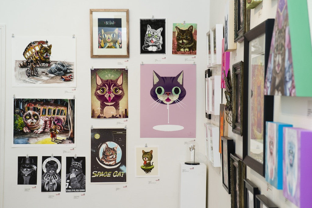 A gallery wall features a vibrant collection of cat-themed art, including a fine art giclee by Casey Weldon. The pieces showcase imaginative styles, such as cosmic designs and a smiling cat on a pink background.