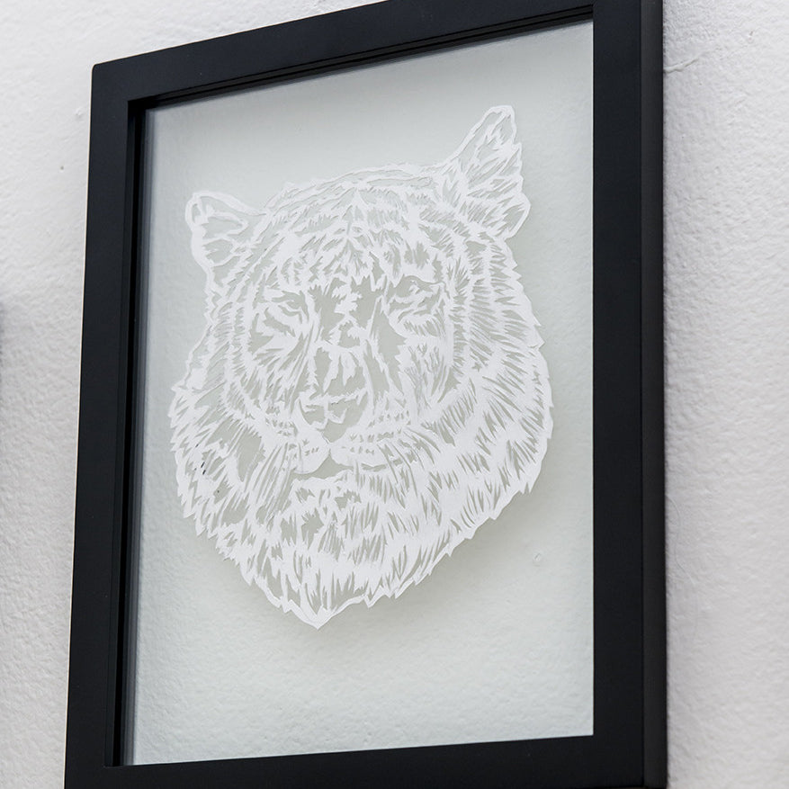 Elaine Penwells framed artwork displays an intricate hand-cut paper design of a tigers head on white. The black frame highlights the detailed fur and facial features, with the Spoke Art logo in the corner, inspired by 1984 artistry.