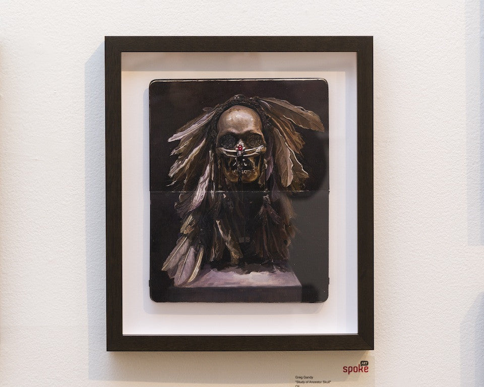 A framed artwork by Greg Gandy features an Ancestor Skull with feathers and beads against a dark background. The black frame with a subtle white border enhances this evocative piece, displayed on a white wall.