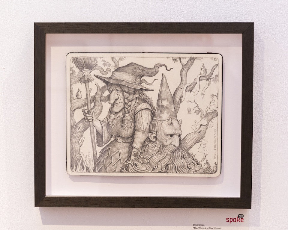 The Brun Croes framed black-and-white illustration features a witch with a broom and a wizard with a beard and pointed hat, surrounded by branches and mystical elements. The detailed graphite pencil artwork hangs elegantly on a white gallery wall.