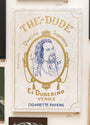 A Casey Weldon-inspired art poster titled The Dude showcases a bearded man with a card and cigarette. Styled as a vintage rolling paper pack, it features Qualite Superieure, El Duderino, and Cigarette Papers.