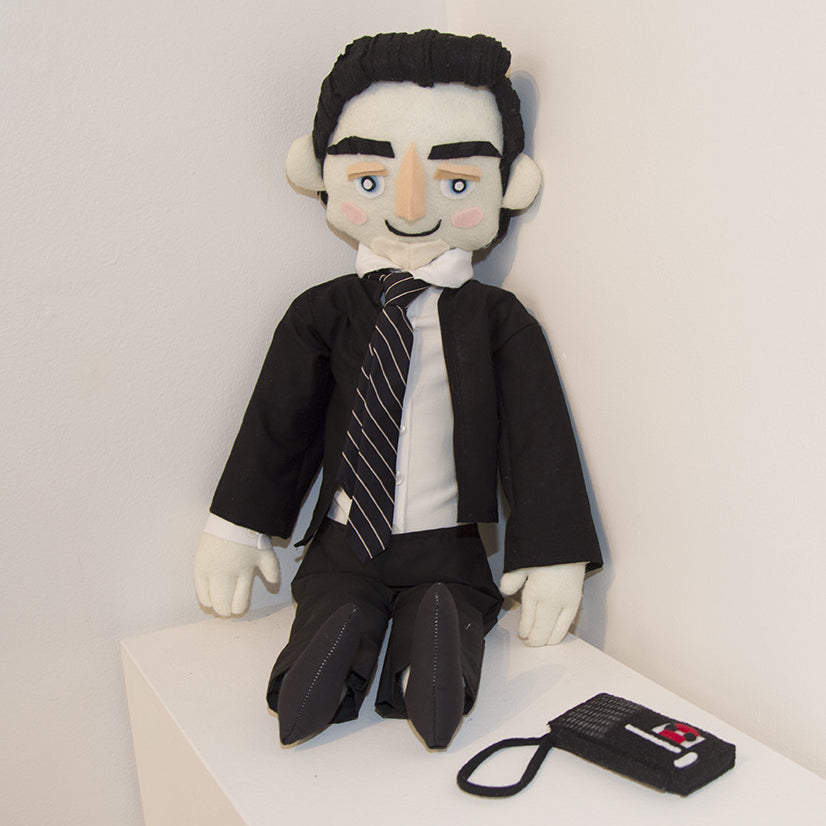A plush FBI Special Agent doll with black hair, a suit, tie, and shoes leans on a white wall. Displaying Tessa Morrisons exquisite fiber art with a small smile, it sits near a small black and red device with a strap.