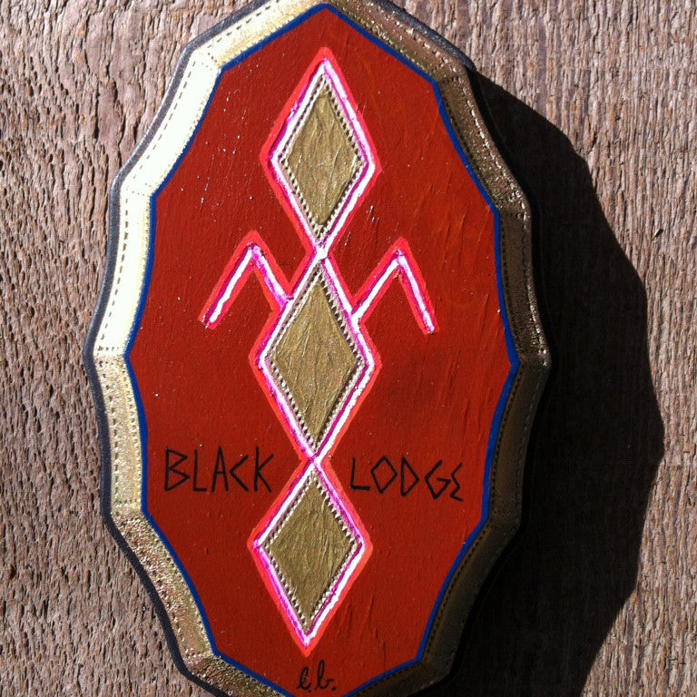 A red emblem displays gold and pink diamond motifs and lines, with the text Black Lodge by Evan Harris artistically inscribed below, all set against a luxurious wooden backdrop.
