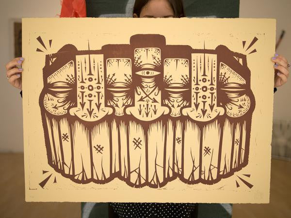 A person holds a stunning GATS artwork with symmetrical abstract humanoid figures and geometric patterns in brown on a beige background, crafted as a limited edition of 80 using the linoleum cut technique.