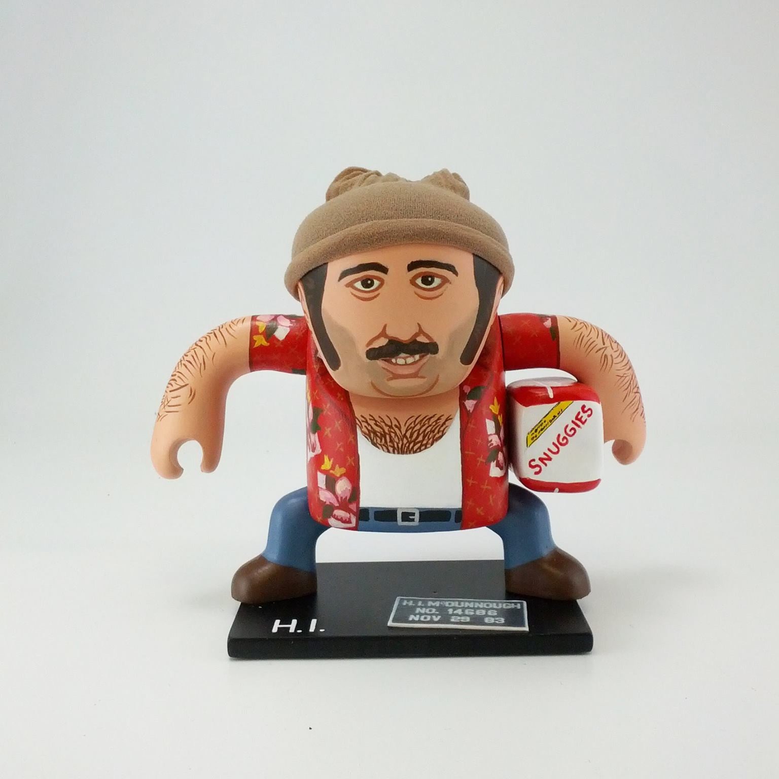 A cartoon figure of Geoff Trapp sports a mustache, red floral shirt, brown pants, beige beanie, and holds a Smugglers mug. He stands atop a black base labeled H.I. against a white background, combining mixed media elements.