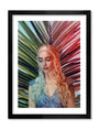In this Stormborn collection archival pigment print by Kemi Mai, a woman with long, wavy silver hair stands before a vibrant, multicolored palm leaf backdrop. She wears a light blue dress and gazes downward serenely. The image is beautifully framed in black.