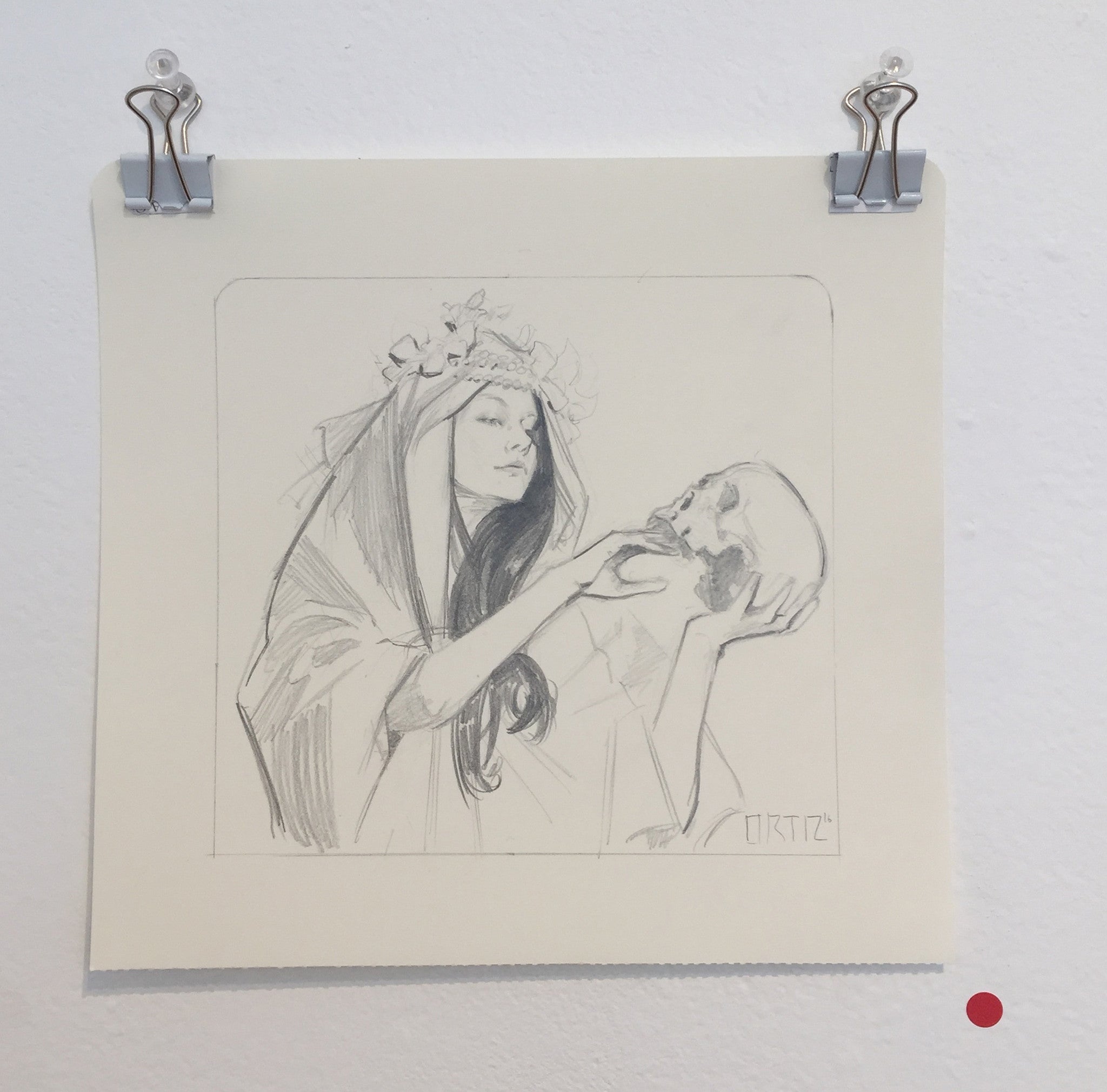A Karla Ortiz ink and graphite sketch shows a person in flowing garments with a floral headpiece holding a skull. The artwork, mounted on the wall with clips, includes a small red dot on the lower right side.