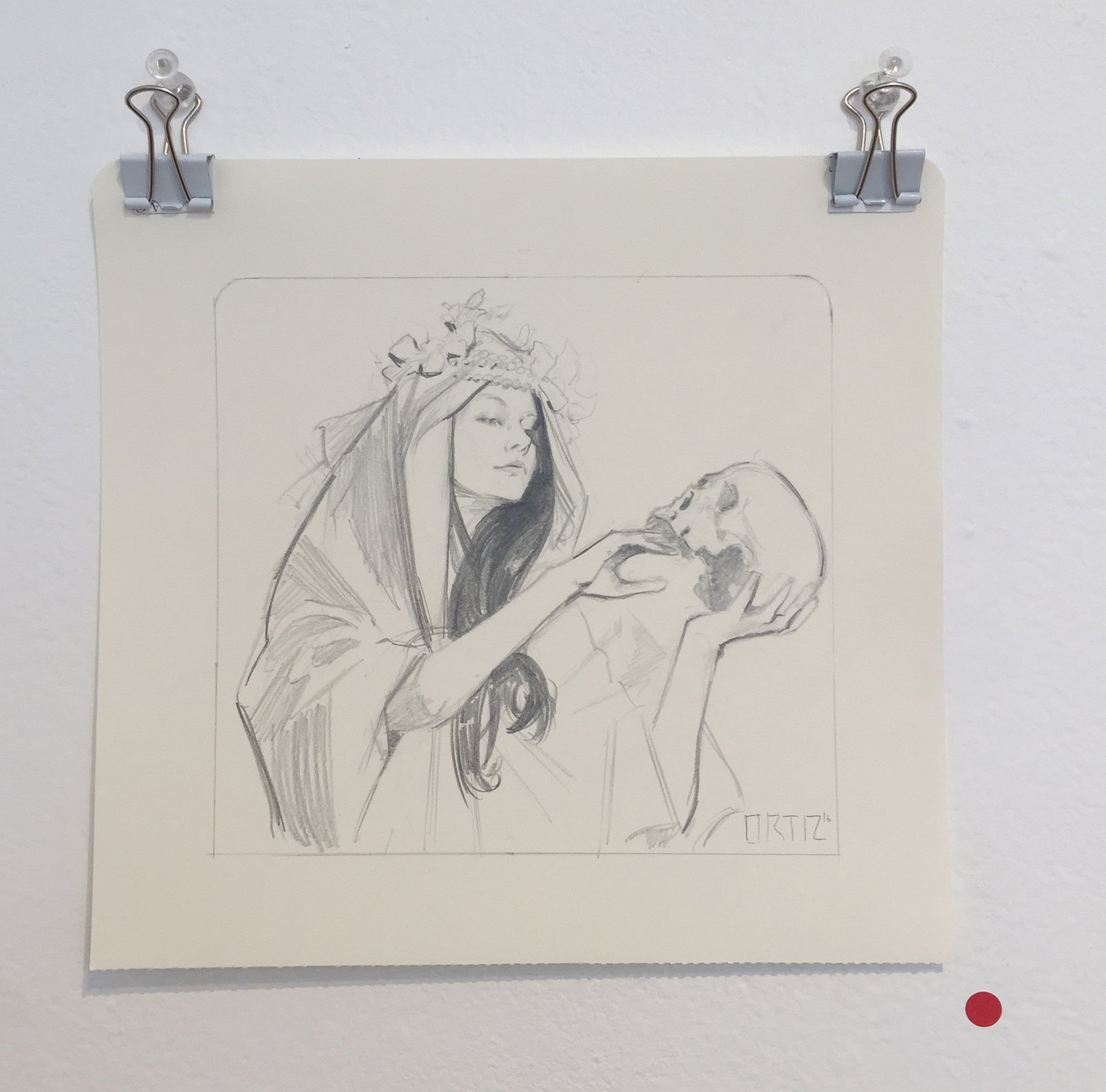A Karla Ortiz ink and graphite sketch shows a person in flowing garments with a floral headpiece holding a skull. The artwork, mounted on the wall with clips, includes a small red dot on the lower right side.