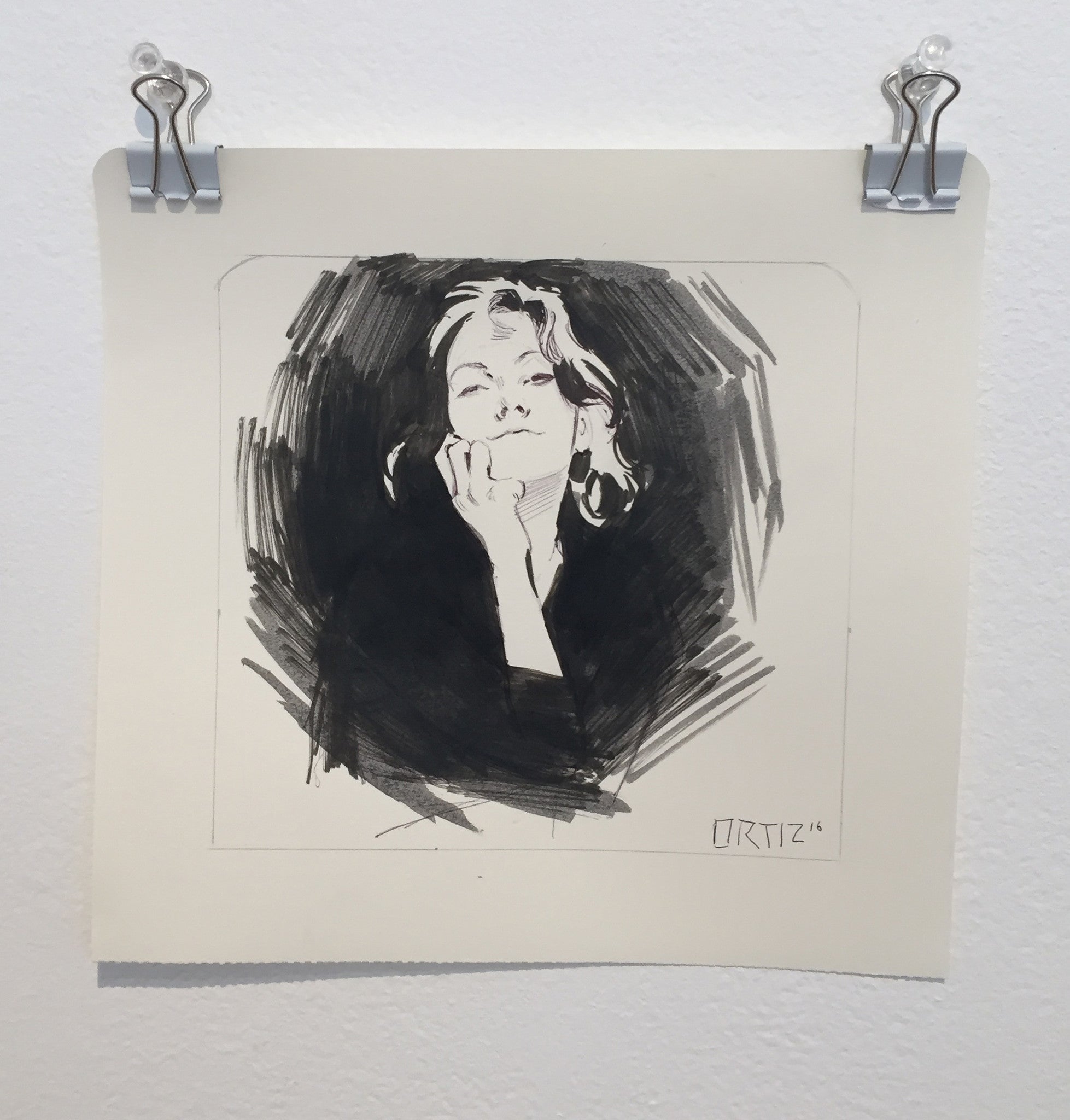Los Bocetos 5 by Karla Ortiz is an ink and graphite sketch on paper, depicting a person with closed eyes and a serene expression, resting their chin on their hand. It hangs elegantly from metal clips on a pristine white wall.