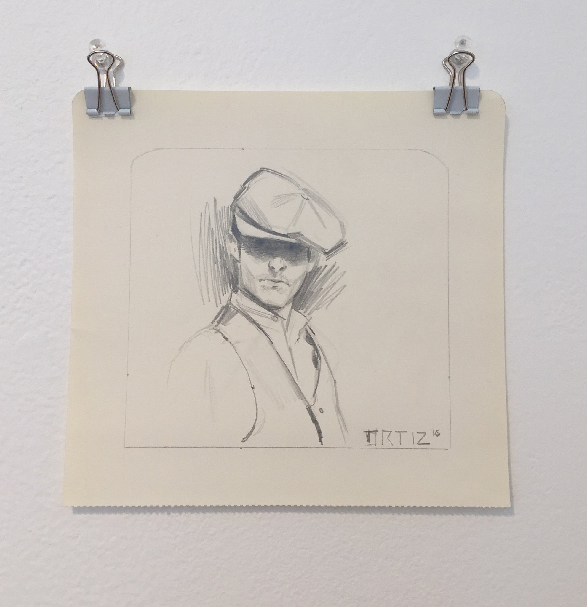 A pencil drawing of a person in a flat cap and jacket, clipped to a wall. Made with ink and graphite, it’s signed ORTIZ 16 in the lower right corner and is from Karla Ortizs Los Bocetos 9 series.