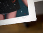 Close-up of Ana Bagayans limited edition fine art giclee print with rough edges on a table. The image shows a figure in a starry outfit, featuring Ana Bagayans white signature in the corner and another smaller red signature near the edge.