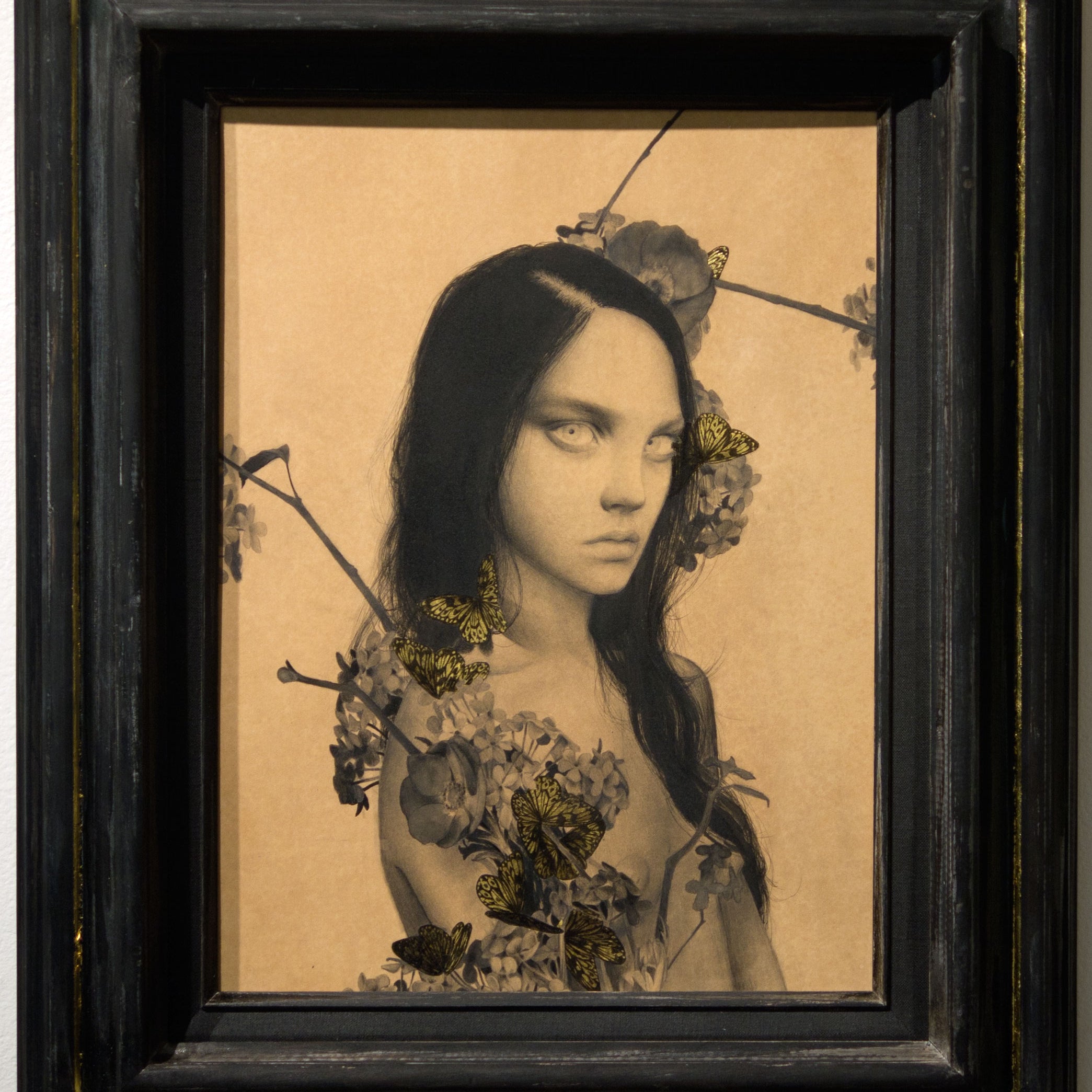 A framed artwork by Alessandra Maria features a solemn figure with long dark hair, surrounded by flowers and butterflies on a neutral background. The blend of realistic and surreal elements enhances its enigmatic mood, reminiscent of pieces at Spoke Art.
