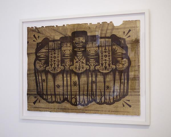 GATS offers framed artwork showcasing a symmetrical design with four abstract faces on a textured brown background, reminiscent of a linoleum cut. It features intricate patterns and a central eye motif for added depth and intrigue.