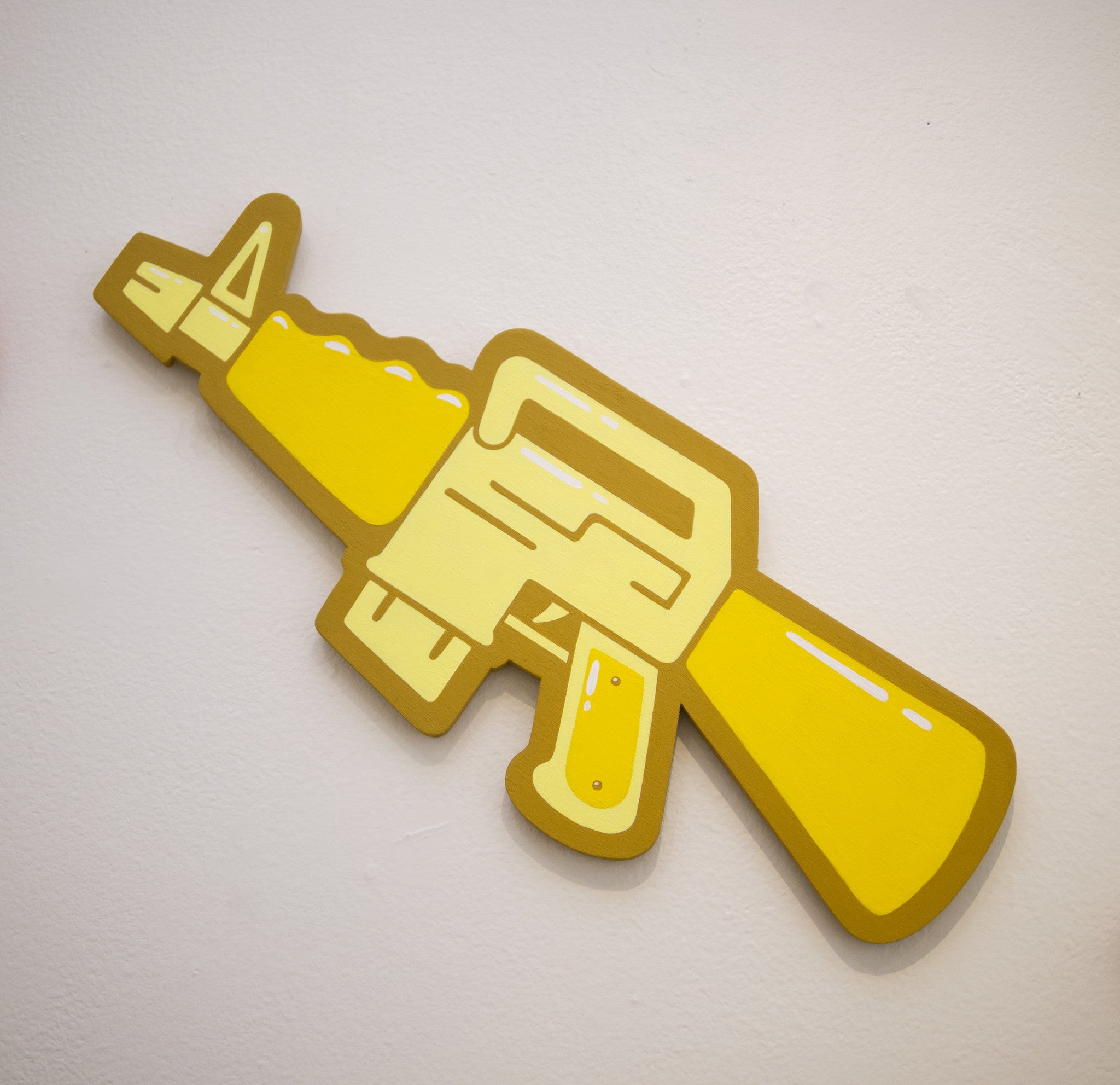 A yellow, cartoonish 3D cutout of an assault rifle by Matt Ritchie is mounted on a white wall, featuring bold outlines and a playful, graphic design reminiscent of hand-cut wood art.