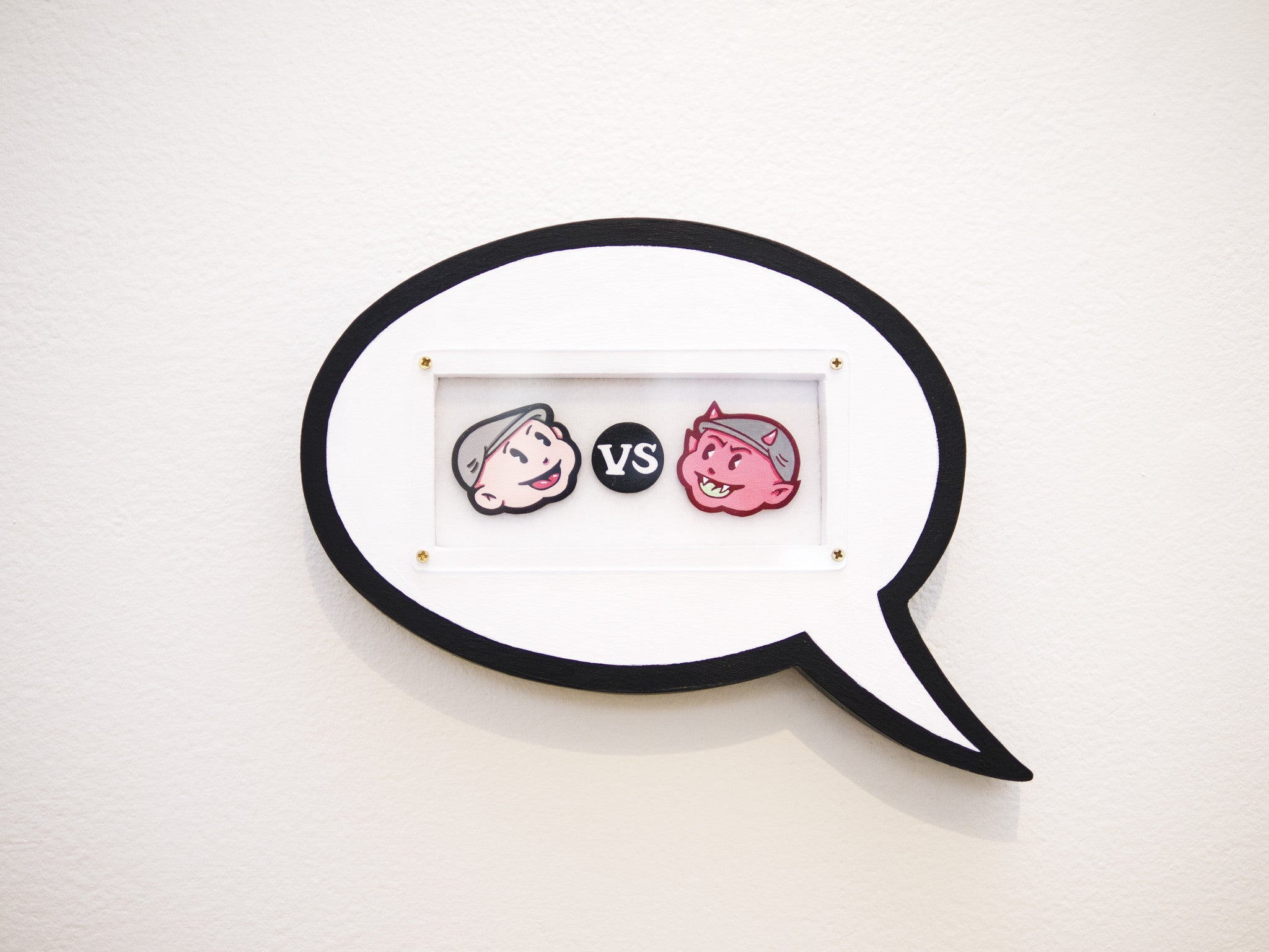 An acrylic art piece inspired by Matt Ritchie features two cartoon faces—a smiling boy on the left and a smiling pink devil on the right—inside a speech bubble frame with vs between them, set against a plain white wall.