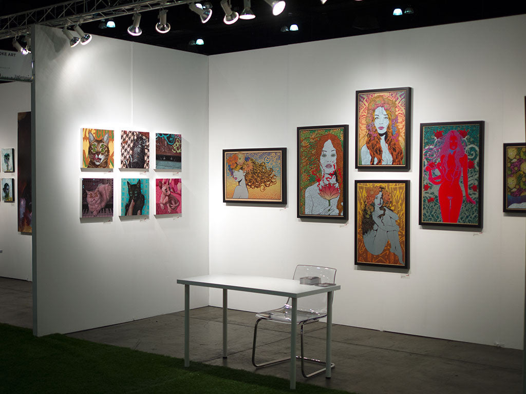 An art gallery with white walls displays vibrant framed artworks, including Casey Weldons portraits and abstract designs on wood panels. A clear plastic chair and a small white table are illuminated by bright ceiling lights in the center.