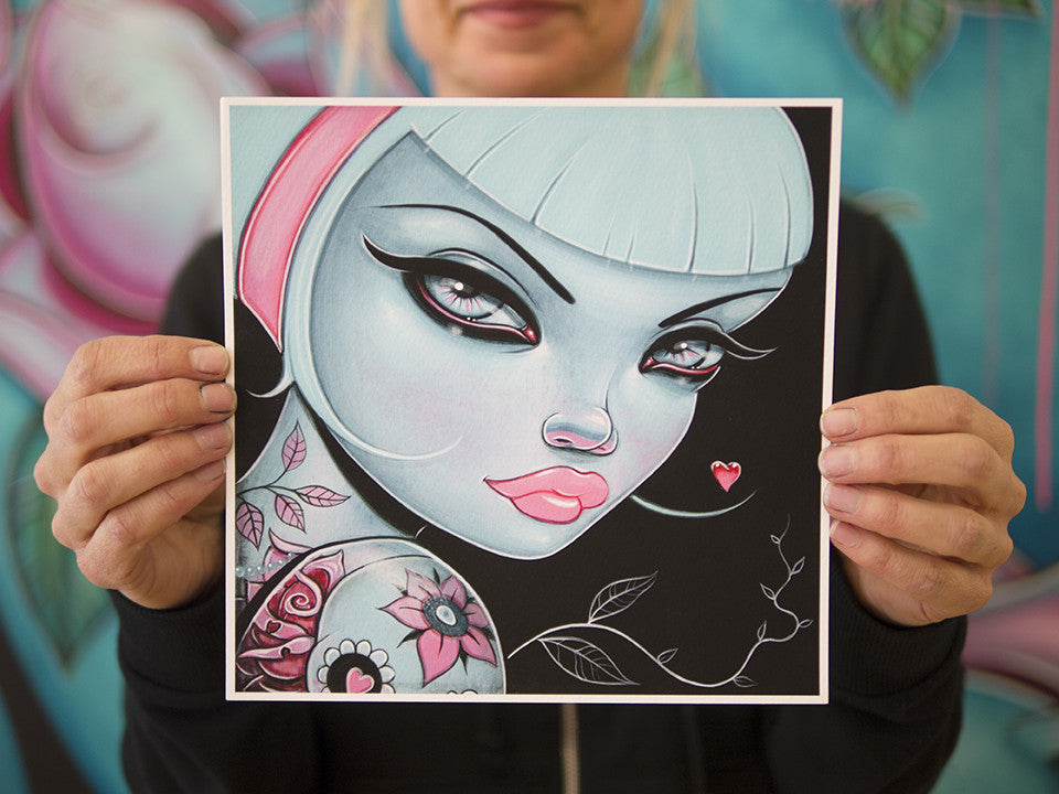 A person holds a vibrant Caia Koopman fine art giclee, featuring a stylized woman with blue skin, sharp eyeliner, pink lips, a bob haircut, floral tattoos, and a small red heart earring. This piece reflects the imaginative style of Mommys Little Monster.