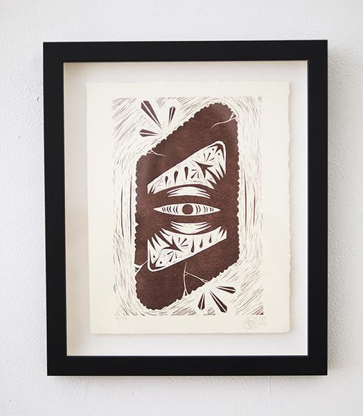 Framed artwork by GATS showcasing Infinite Eye, an abstract eye design in brown tones with geometric patterns on white paper. This letterpress linoleum cut is part of a signed and numbered edition, elegantly displayed against a plain white wall.