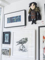 The display evokes a tribute to George R.R. Martin and J.R.R. Tolkien, featuring framed Allison Reimold artwork: a forest scene, a raven on a skull, and a small warrior figure. A fabric doll in medieval clothing adds charm to the white walls bathed in ambient natural light, akin to The Sight at Spoke Art Gallery.