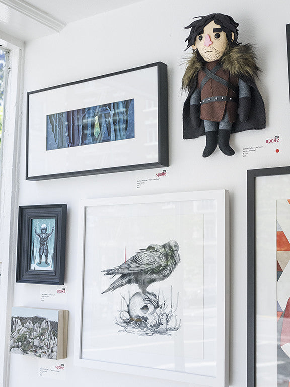 The display evokes a tribute to George R.R. Martin and J.R.R. Tolkien, featuring framed Allison Reimold artwork: a forest scene, a raven on a skull, and a small warrior figure. A fabric doll in medieval clothing adds charm to the white walls bathed in ambient natural light, akin to The Sight at Spoke Art Gallery.