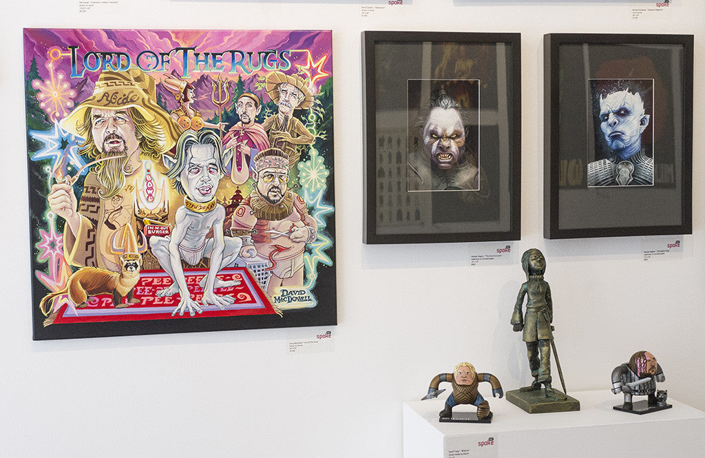 A gallery wall at Spoke Art exhibits a humorous Lord of the Rugs parody by Dave MacDowell with vivid caricatures, next to two framed portraits with dark expressions. Below, a shelf holds small figurines that mimic characters with exaggerated features.