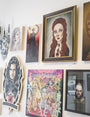 A wall at Spoke Art gallery features diverse artworks like Sandi Calistro portraits and a Melisandre-inspired fantasy collage, with styles from realistic to cartoonish, vibrant colors, and dark tones.