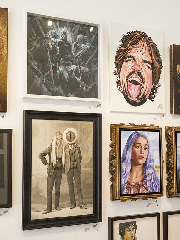A gallery wall at Spoke Art showcases diverse works, such as Eric Bonhomme’s fantasy scene Forever Winter with dark figures, a portrait of a person shouting, a sepia image of two figures, and a vibrant portrayal of someone with long purple hair.