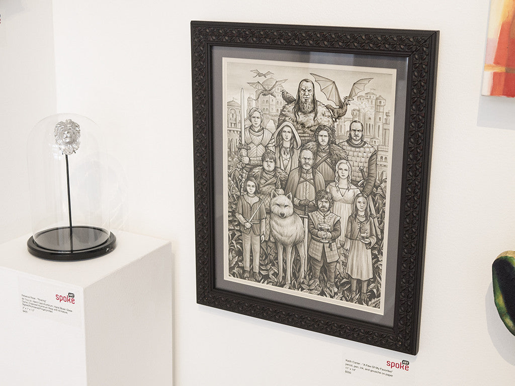 A black and white fantasy drawing with a dragon and undead figures complements the roaring wearable ring by Rebecca Rose on display. The framed art hangs on a gallery wall next to a hand-blown glass dome, with its price tag beneath.