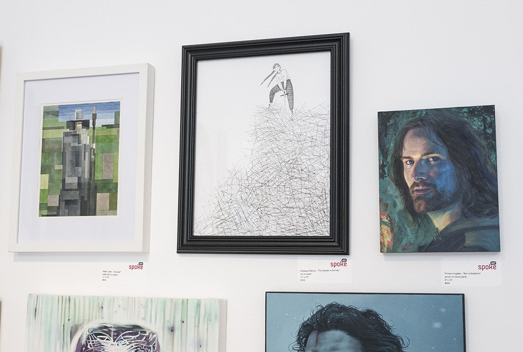 In the Spoke Art gallery, a wall displays framed artworks: left, a geometric abstract; center, a figure amid scribbles; right, Chelsea OByrnes detailed portrait of a man with shoulder-length hair. Small labels accompany each piece.