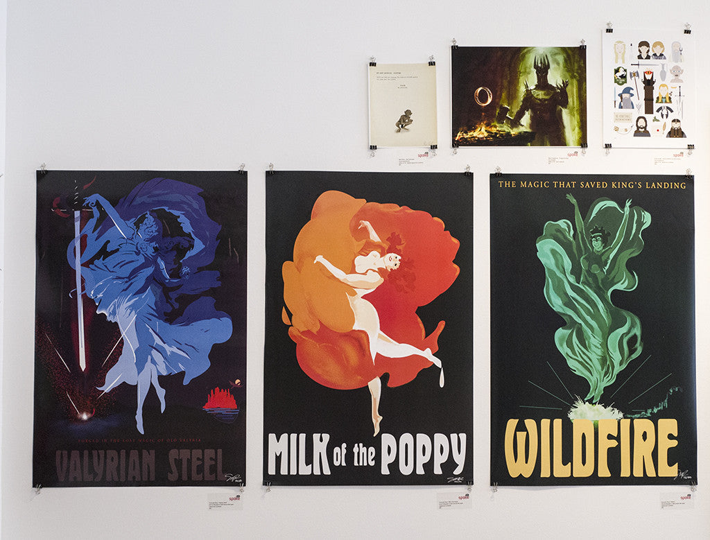 Three fine art lithograph posters by Fernando Reza decorate the wall with a fantasy theme: Valyrian Steel shows a knight and sword, Milk of the Poppy features a dancing figure, and Wildfire displays a green spectral form. All are custom framed with additional small artwork above.