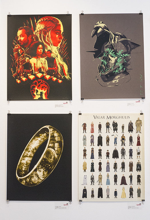 Four fantasy-themed posters by Joshua Budich are displayed on a wall, featuring screen prints of characters and items from different fictional series. They include dragons from House Targaryen, a ring, and various stylized figures, each with distinct artwork and colors.