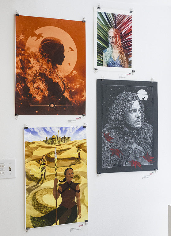 A wall showcases four fantasy-themed posters by Kemi Mai: top left, a Stormborn silhouette with fiery details; top right, vibrant foliage in archival pigment print; bottom left, a desert wanderer; and bottom right, a winter entity amidst snow—a captivating group exhibition.