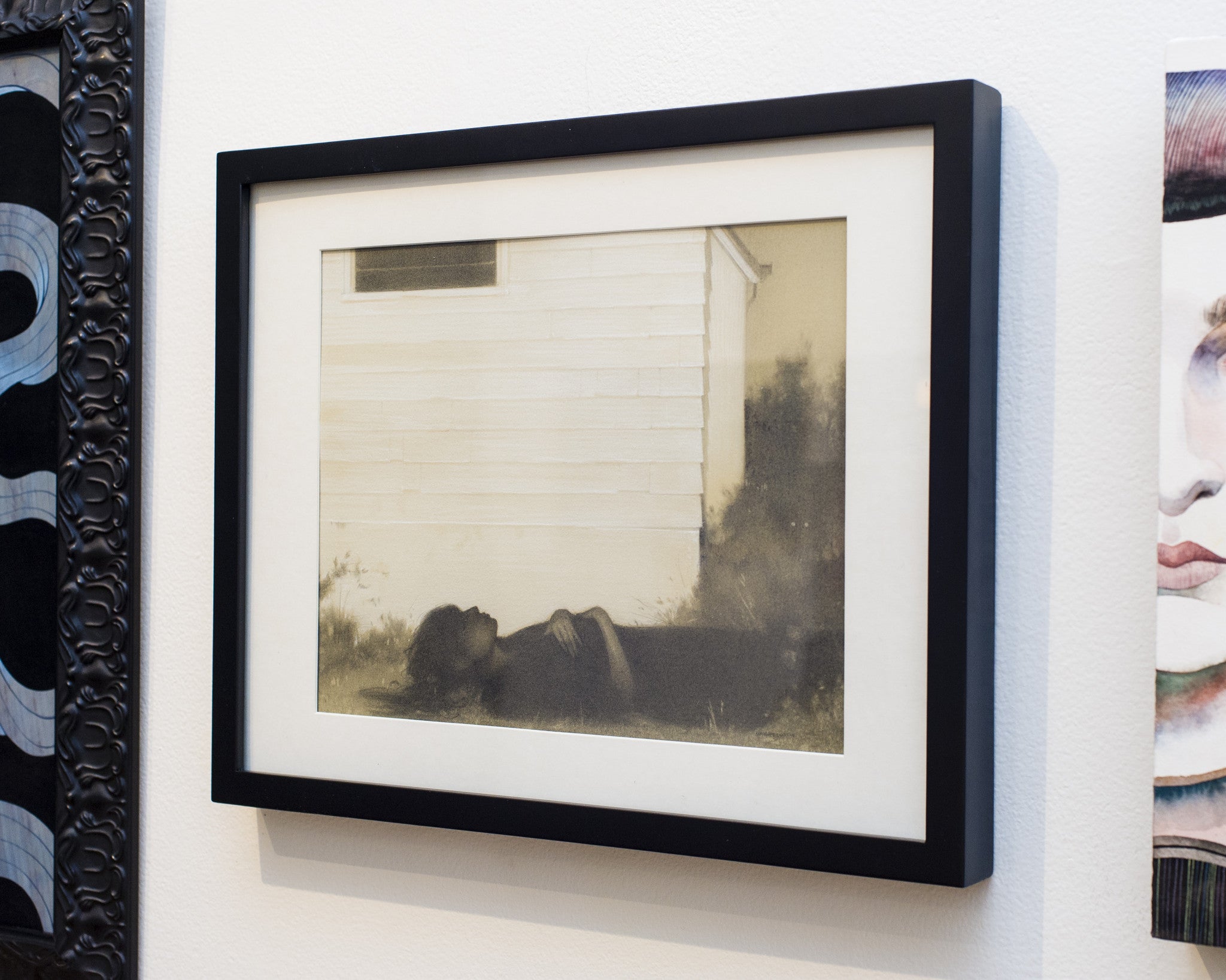 A framed artwork using white charcoal, showing a person by a light building, adds intrigue to a gallery display reminiscent of Sam Wolfe Connellys atmospheric style.