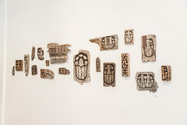 GATS presents an artistic wall display featuring uniquely carved wooden pieces of various shapes and sizes. The pieces resemble burnt driftwood and showcase intricate patterns, including faces and geometric shapes, forming a mosaic-like artwork on a white wall.