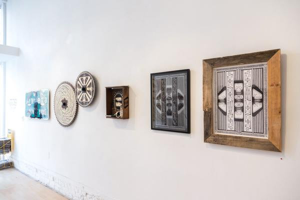 A gallery wall features five GATS art pieces: a canvas painting, two circular weavings, a mask in a wooden box, and custom framed black-and-white geometric artworks. The bright setting with smooth white walls and light wooden floors enhances this unique illustration collection.