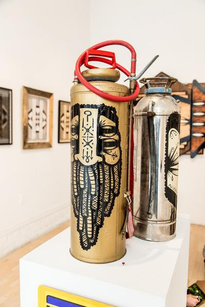 Two vintage GATS fire extinguishers are displayed in an art gallery. The front one is gold with black designs and a red hose; the back one is enamel silver with a unique pattern. Framed art pieces adorn the background walls.