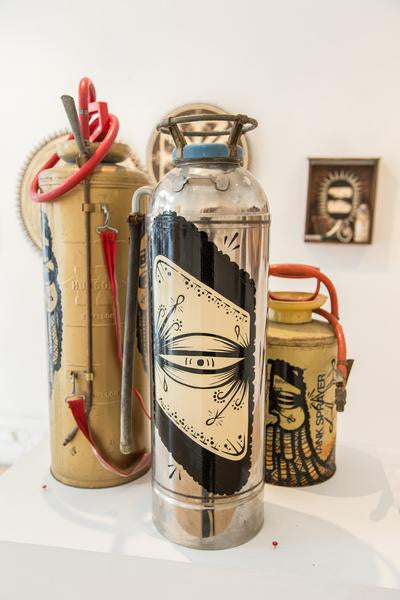 Three vintage fire extinguishers with unique enamel-painted designs are showcased. The centerpiece features GATSs signature stylized eye pattern. Red hoses drape over them, and a small framed artwork is visible behind them.