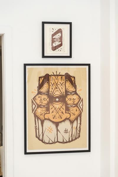Two framed artworks adorn the white wall. The larger GATS piece, a signed and numbered edition, features an abstract face with geometric patterns in brown and beige. Above it, a smaller frame displays two stylized feathers on a brown background, echoing an Infinite Eye design.