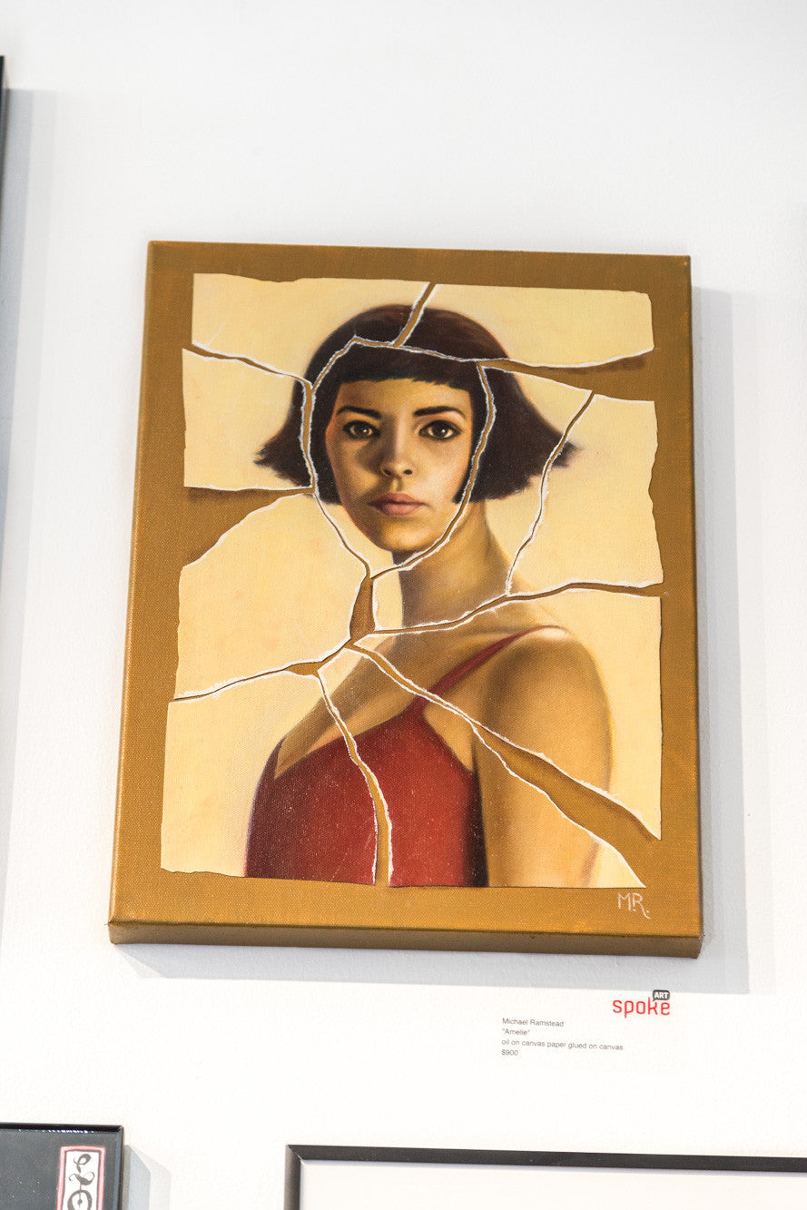 A wall displays a fragmented painting by Michael Ramstead of a woman in a red top with short dark hair, reminiscent of Amelie. Cracks throughout resemble a shattered surface, and it bears the artists initials MR at the bottom right.
