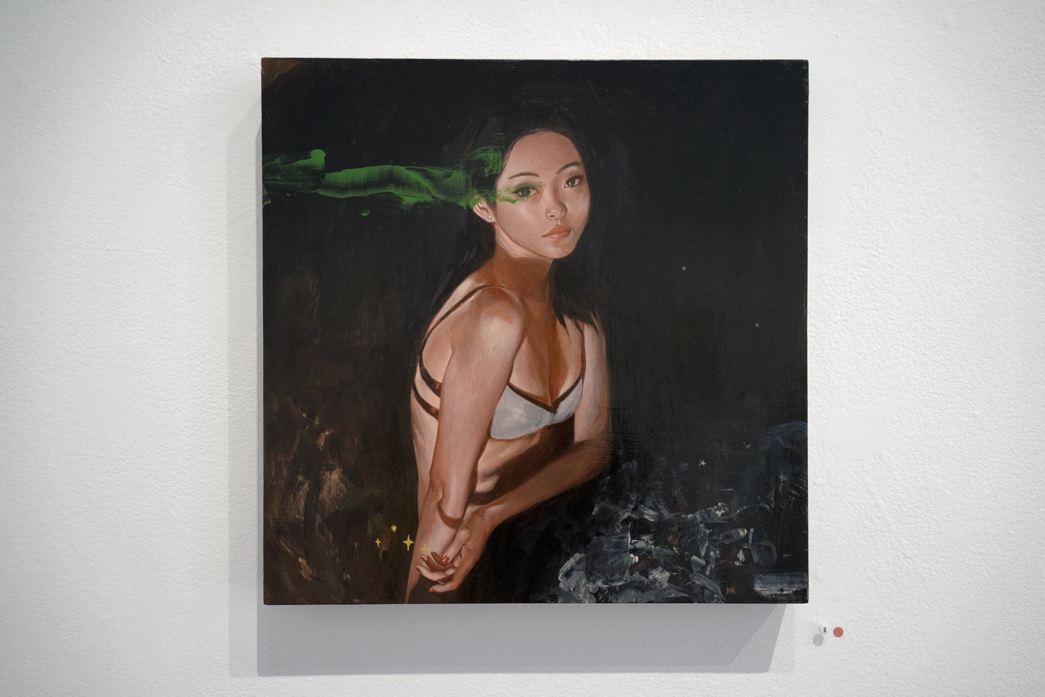 A Helice Wen painting showcases a woman in a bra with crossed arms against a dark backdrop, highlighted by a green brushstroke near her face evoking Northern Light. This acrylic on wood panel features abstract, smoky textures and is displayed on a white wall.