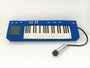 Steff Bombs blue felt toy keyboard, inspired by Bobs Burgers art, features black and white keys, control buttons, and a display. A black cord connects it to a silver and black felt microphone, all set for Gallery1988 fans on a white surface.