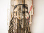 The GATS rustic artwork on burnt driftwood displays a stylized face with closed eyes, intricate patterns, and dark lines. It exudes a weathered look with red paint traces, evoking the mysterious Drifting Forest as it leans against a white wall.