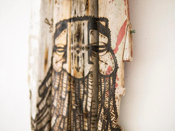 The GATS rustic artwork on burnt driftwood displays a stylized face with closed eyes, intricate patterns, and dark lines. It exudes a weathered look with red paint traces, evoking the mysterious Drifting Forest as it leans against a white wall.