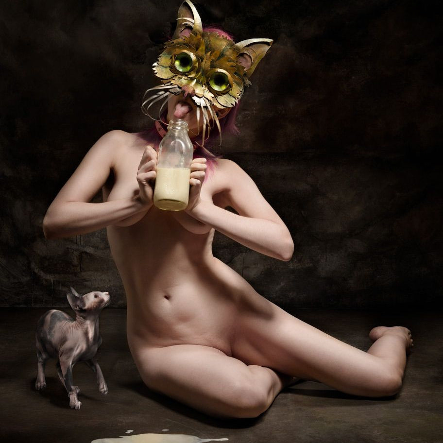 In a dim room with a textured backdrop, someone kneels wearing a cat mask and drinks milk as a small hairless cat watches. This intriguing scene echoes surreal moments from Ransom & Mitchells exclusive posters for the Lil Bub Art Exhibit.