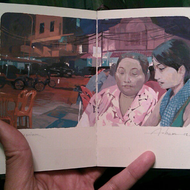 A hand holds the sketchbook open, showing Andrew Hems gouache painting titled The Visitor, depicting two people together at night amid buildings, lights, and empty chairs.