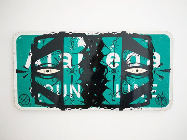 GATS street sign artwork showcases stylized black eyes and abstract patterns on a green and white background, crafted with enamel. Bold black shapes and intricate details offer a mysterious, artistic touch at the Alameda County Line.