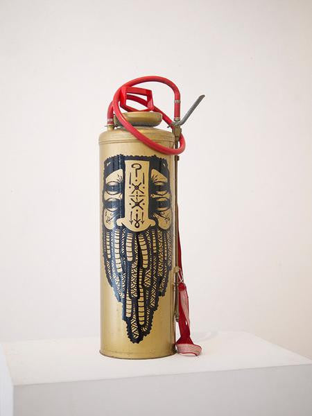 A vintage GATS fire extinguisher, featuring intricate enamel artwork of a stylized face and beard in gold, rests on a white pedestal. Its red hose is elegantly coiled on top against a plain background.