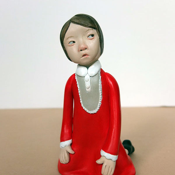 A hand-sculpted resin figurine by Doubleparlour features a child with short hair kneeling in a bright red dress with a white collar and cuffs, detailed with acrylic paint. The child’s thoughtful expression is contrasted against a plain, light background.